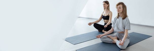 Two athletic attractive girls sit in lotus position on yoga mat indoors .Yoga, fitness and healthy lifestyle.  - Foto, afbeelding
