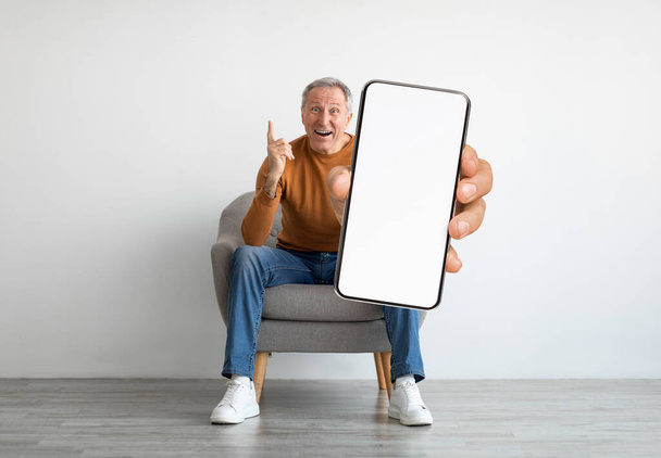 Great Offer. Excited Senior Man Sitting In Chair And Holding Big Blank Smartphone, Elderly Male Demonstrating Huge Mobile Phone With White Screen At Camera, Advertising New App, Collage, Mockup - Фото, зображення