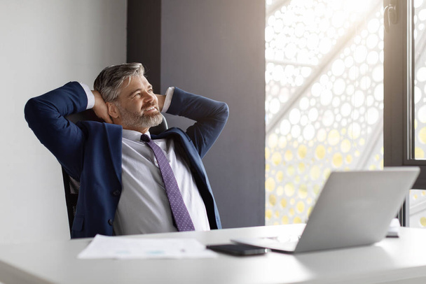 Portrait of happy mature male entrepreneur relaxing at workplace in office, smiling glad middle aged businessman in suit sitting at desk and leaning back in chair with hands behind head, free space - 写真・画像