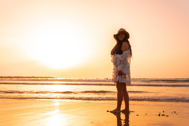 Portrait of a woman at sunset in a white dress with a hat walking by the sea at low tide - Photo, image