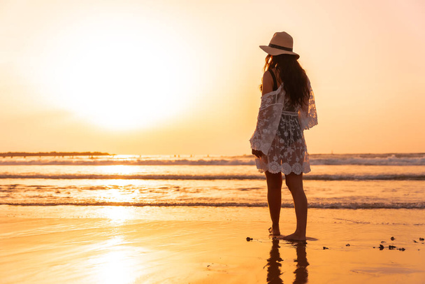 A woman at sunset in a white dress with a hat walking by the sea at low tide - Photo, Image