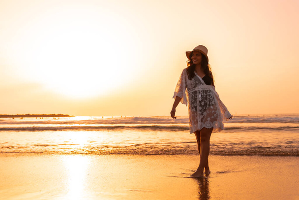 Egy nő a naplementében, fehér ruhában, kalappal a tengerparton sétálva. - Fotó, kép