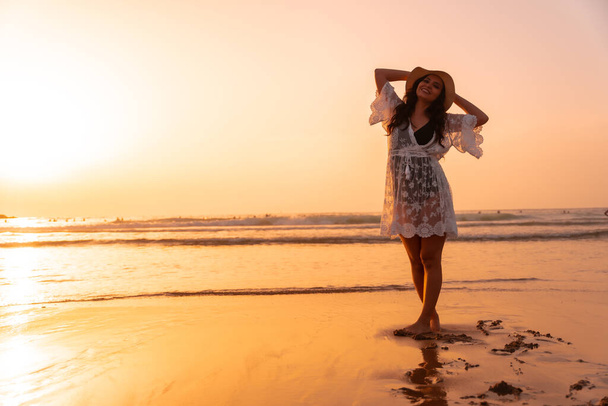 A woman in the sunset in a white dress at the sea in summer with a hat - Foto, Bild