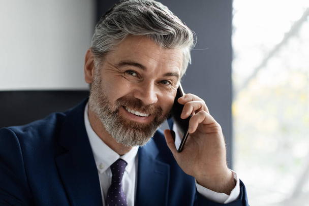 Obchodní hovor. Detailní záběr usměvavý zralý podnikatel mluví na mobilním telefonu v kanceláři, hezký středověký muž podnikatel nosí oblek sedí na pracovišti a po rozhovoru na mobilním telefonu - Fotografie, Obrázek