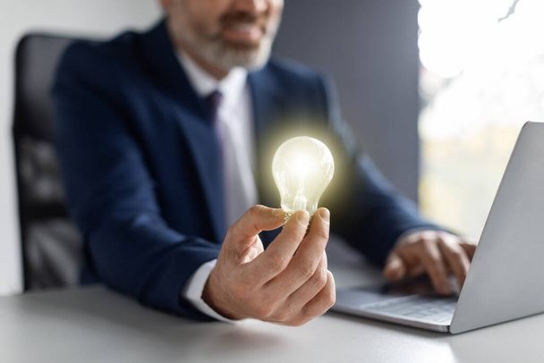 Business Innovations. Middle Aged Businessman Holding Illuminated Light Bulb While Sitting At Workplace With Laptop Computer, Entrepreneur In Suit Having Startup Idea, Closeup Shot, Cropped - Foto, immagini