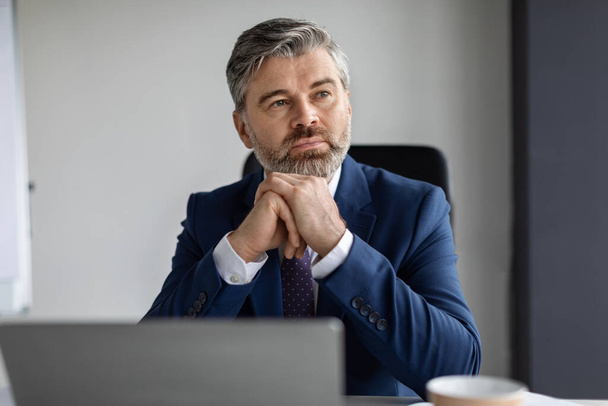 Portrait Of Thoughtful Middle Aged Male Entrepreneur Sitting At Desk In Office And Looking Away, Pensive Mature Businessman Wearing Suit Thinking About Business Strategy And New Ideas, Closeup - Фото, зображення