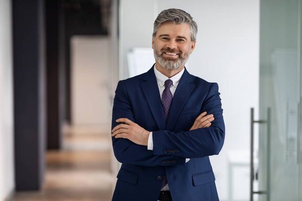 Portrait Of Confident Handsome Middle Aged Businessman In Suit Standing With Folded Arms, Successful Male Entrepreneur Posing In Office Hall, Advertising Professional Services, Smiling At Camera - 写真・画像