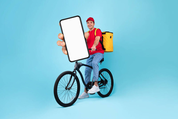 Food Delivery App. Young Courier Guy On Bicycle Showing Big Blank Smartphone With White Screen At Camera, Smiling Deliveryman In Uniform Recommending Mobile Application For Online Order, Mockup - Zdjęcie, obraz