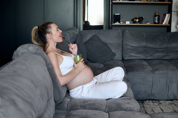 pregnant woman eating a smoothie in the living room. Healthy pregnant eating lifestyle concept - 写真・画像