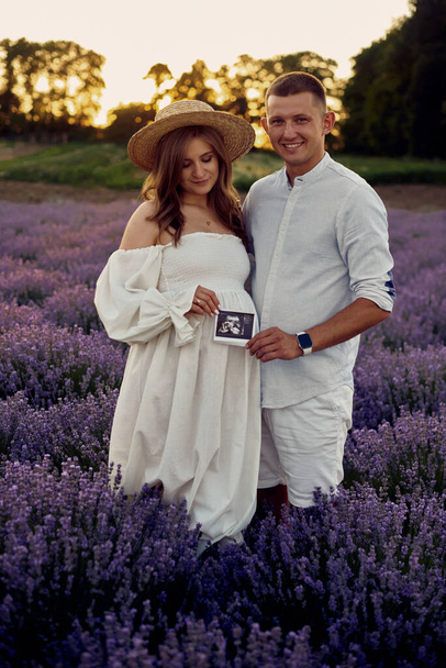 portrait of a young beautiful pregnant couple on a lavender field at sunset holding first ultrasonography image. Happy family concept - Foto, imagen
