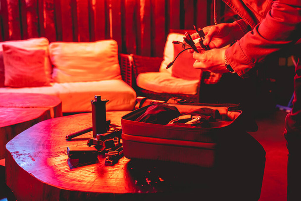 Hands of photographer preparing photo accessories, batteries and cables from his bag in a rustic wood table under colorful red and purple light in the night - Photo, image