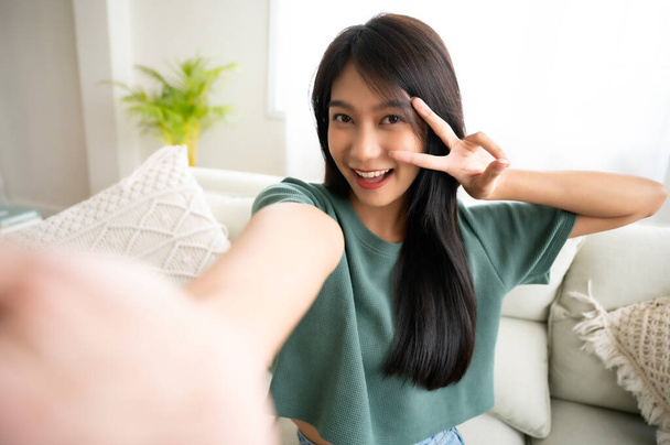 Pretty young asian female with big smile sitting at living room. She having fun taking light cheerful selfie on blurred background - Фото, зображення