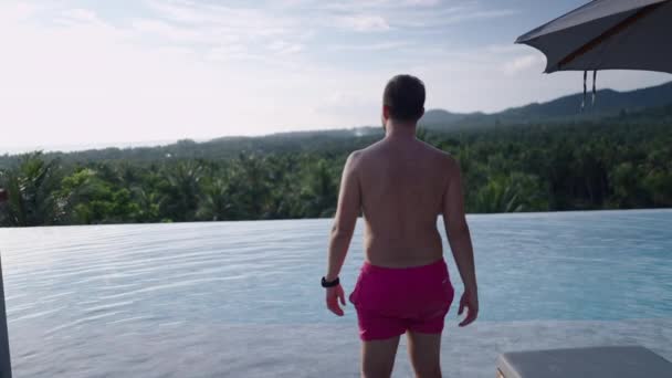 A shirtless caucasian man walks up to the swimming pool in swim trunks to see a picturesque jungle forest panorama from the hotel lounge at sunset - slow motion - 4K Horizontal video - Filmati, video