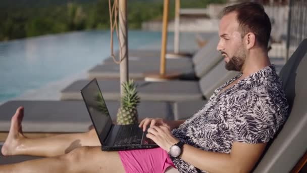 Giovane imprenditore che lavora durante le vacanze, utilizzando un computer portatile e rispondendo alle telefonate mentre si trova su una sdraio a bordo piscina in un resort tropicale - 4K Video orizzontale - Filmati, video