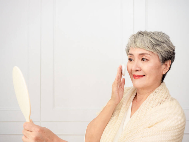Portrait of mature aged asian woman 50s touching face skin looking in mirror reflection. Smiling mature old lady pampering, healthy moisturized skin care, aging beauty, skincare treatment cosmetics concept. - Photo, Image