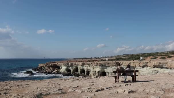 Time-lapse of a young couple from behind is sitting on the bench and looking at sea caves near cape Greco in a national park with turquoise water sea in Ayia Napa, Cyprus.  - Footage, Video