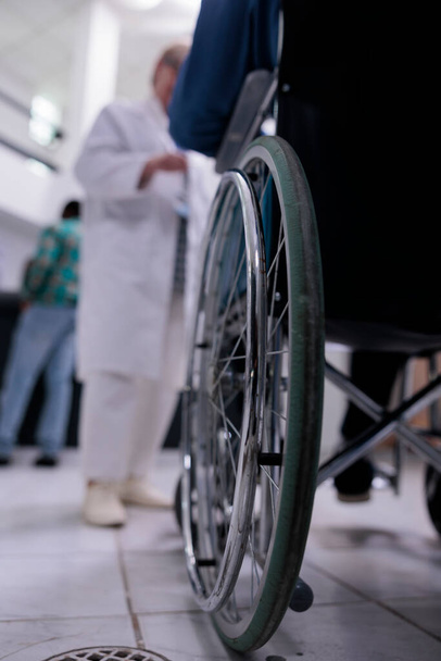 Gros plan d'un aîné vivant avec un handicap utilisant un fauteuil roulant à la réception d'une clinique privée parlant avec un médecin au sujet d'un rendez-vous. Focus sélectif sur la roue en fauteuil roulant dans le hall de l'hôpital occupé. - Photo, image
