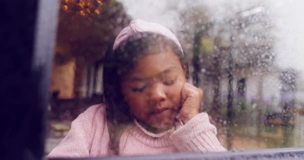 Bored female child looking outside on a wet winter day. Unhappy child feeling lonely and depressed while looking at the bad weather outdoors. Sad little girl standing by the window on a rainy day - Video, Çekim