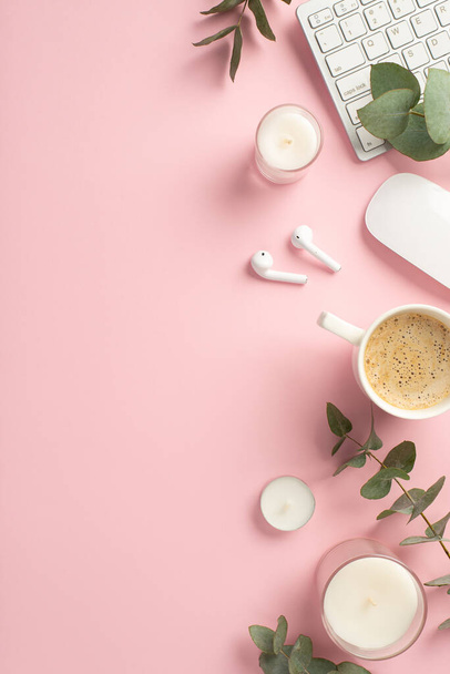 Business concept. Top view vertical photo of workspace keyboard candles computer mouse cup of coffee wireless earbuds and eucalyptus on isolated pink background with copyspace - 写真・画像