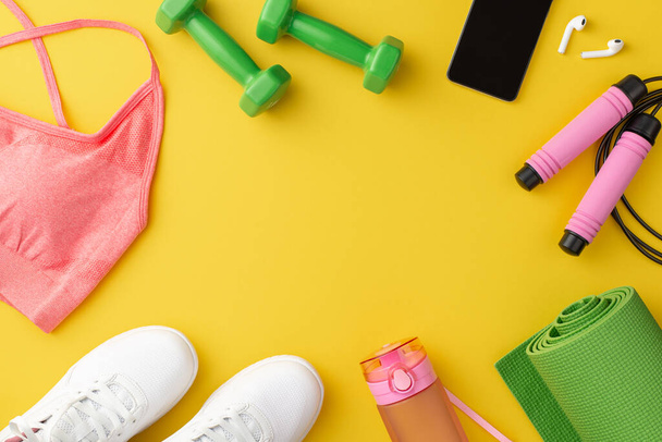 Fitness concept. Top view photo of pink sports top white sneakers skipping rope bottle dumbbells yoga mat earbuds and smartphone on isolated yellow background with copyspace in the middle - 写真・画像