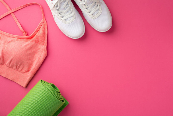 Fitness-Zubehör-Konzept. Foto von oben von der grünen Yogamatte rosa Sporttop und weiße Schuhe auf isoliertem rosa Hintergrund mit Kopierraum - Foto, Bild