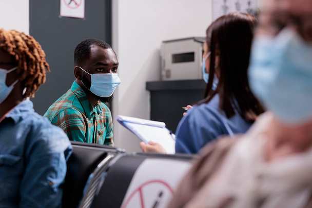 Male patient with face mask talking to assistant, sitting in waiting area at hospital reception lobby. Man doing healthcare consultation with medical worker, nurse and person at facility. - Fotó, kép