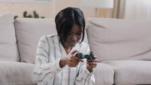 Young focused happy african american woman sitting in living room playing console uses controller plays video game resting at home enjoying playtime alone indoors having fun leisure time hobby concept - Imágenes, Vídeo