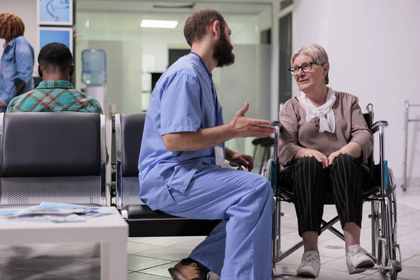 Male nurse consulting woman with chronic disability in hospital waiting area at medical facility. Patient wheelchair user with impairment talking to specialist at healthcare clinic. - Foto, Imagen