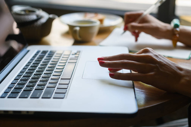 Close-up van de hand van een vrouw die werkt met een laptop in een koffieshop. Taart, laptop, theepot liggen op tafel.. - Foto, afbeelding