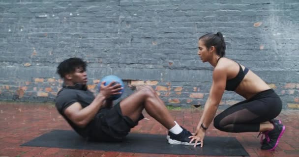 Vidéo 4k d'une jeune femme sportive tenant un jeune homme athlétique pieds pendant que nous faisons des rebondissements assis avec un ballon de médecine au ralenti tout en travaillant à l'extérieur sous la pluie. - Séquence, vidéo