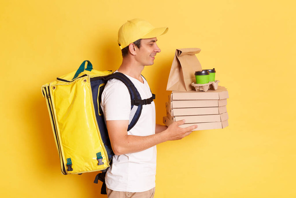 Side view portrait of man courier with thermo backpack in white T-shirt and cap isolated over yellow background, giving online order to client, bringing pizza, coffee and parcel with food. - Foto, Imagem