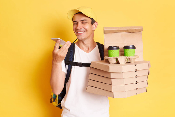 Smiling man courier in white T-shirt and cap isolated over yellow background, holding pizza and takeaway coffee, recording voice message or using assistant for searching correct address. - Fotoğraf, Görsel