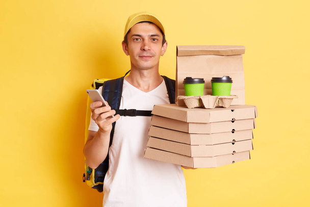 Horizontal shot of serious calm man courier with thermo backpack in white T-shirt and cap, looking at camera, holding client's order, standing with smart phone in hands isolated over yellow background - Photo, image
