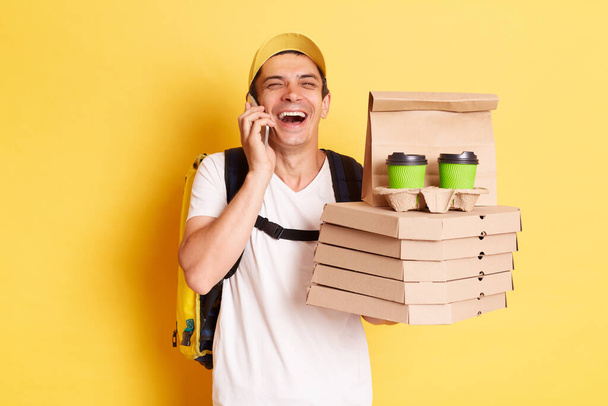 Indoor shot of happy laughing man courier with thermo backpack in white T-shirt and cap isolated over yellow background, talking phone, holding boxes with pizza and takeaway coffee. - Zdjęcie, obraz