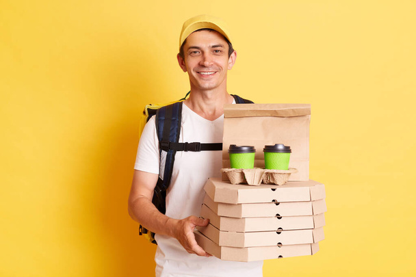 Image of satisfied man courier with thermo backpack in white T-shirt and cap isolated over yellow background, holding boxes with pizza and disposable cups with coffee or tea, delivering order. - Foto, Imagen