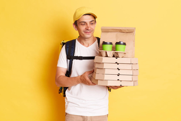 Image of calm smiling deliveryman with thermo backpack in white T-shirt and cap standing against yellow wall, holding and looking at pizza boxes and disposable cups for delivering. - Photo, image