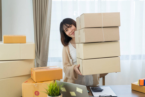 Happy young asian woman startup small business freelance holding parcel box and computer laptop and sitting on chair, Online marketing packing box delivery concept - Фото, изображение