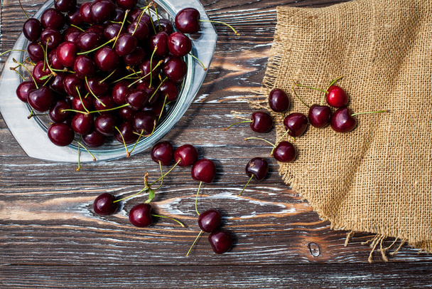 A lot of ripe red cherries on twigs lie in a glass plate on a dark blue wooden table with a striped texture, covered with cloth. - Foto, Bild