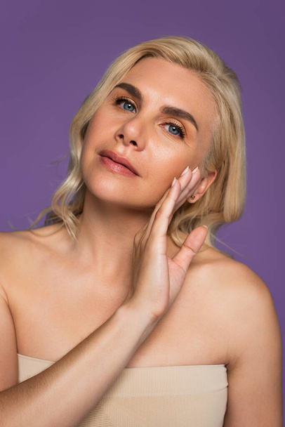 blonde woman with bare shoulders touching cheek and looking at camera isolated on purple - Fotoğraf, Görsel