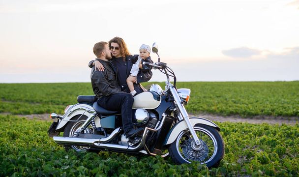 Young beautiful family dad mom and little daughter ride a motorcycle in the field at sunset and pose - Fotó, kép