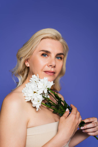 blonde woman with blue eyes looking at camera while holding white flowers isolated on violet - Foto, afbeelding