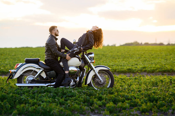 Ein junges schönes Paar, ein Mann und ein Mädchen in Lederkleidung, posieren und umarmen sich auf einem Motorrad auf einem Feld vor dem Hintergrund des Sonnenuntergangs - Foto, Bild