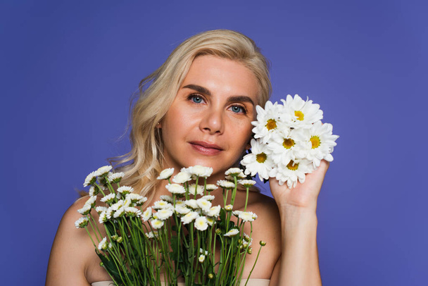blonde woman with bare shoulders holding white flowers and looking at camera isolated on violet - Foto, afbeelding