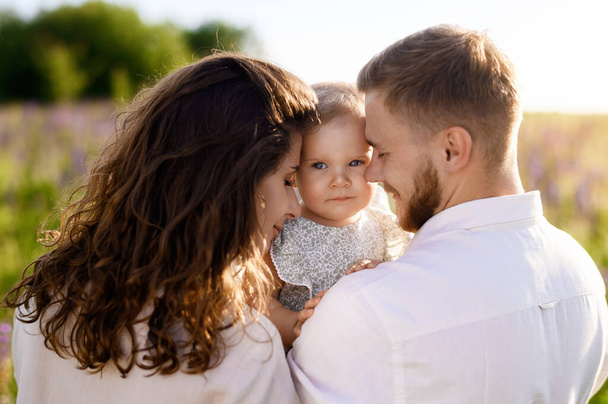 Красивая молодая белая семья папа и мама обнимают маленькую симпатичную девочку на фоне заката в поле. Закрыть - Фото, изображение