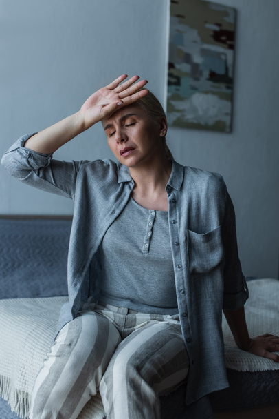 vyčerpaná žena s menopauzou trpí bolestí hlavy při sezení v ložnici - Fotografie, Obrázek
