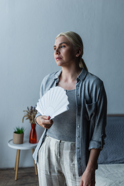 blonde woman with menopause suffering from heat and holding fan in bedroom - Fotoğraf, Görsel