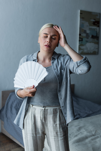 exhausted woman with menopause suffering from heat and holding fan in bedroom - Photo, Image