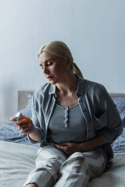 depressed blonde woman with menopause holding bottle with pills and sitting on bed - Foto, Imagem