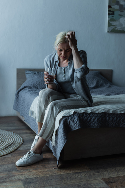 full length of exhausted woman holding glass of water while having migraine during menopause  - Foto, Imagen