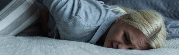 blonde woman lying on bed while feeling pain in stomach during menopause, banner - Foto, afbeelding